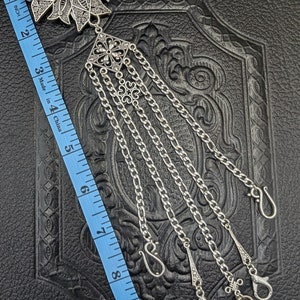 May include: A silver-toned, ornate, floral-shaped pendant with a dark iridescent stone in the center. The pendant is attached to five silver chains with decorative clasps. The chains are approximately 9 inches long.