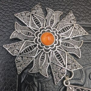 May include: A silver filigree pendant with an orange gemstone in the center. The pendant is shaped like a flower with intricate details.
