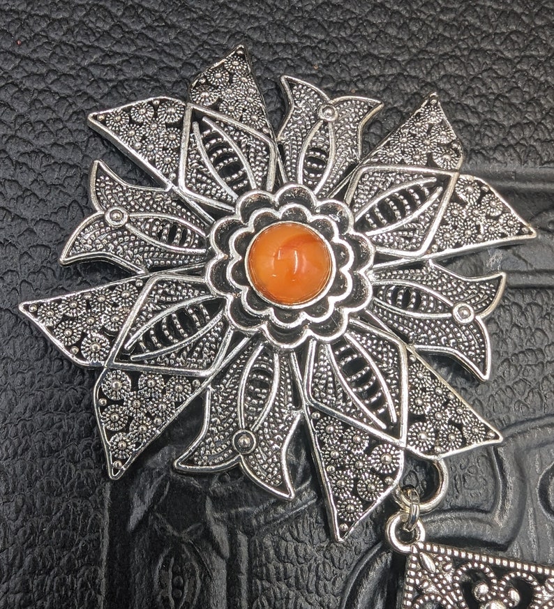 May include: A silver filigree pendant with an orange gemstone in the center. The pendant is shaped like a flower with intricate details.