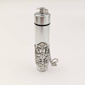 May include: A silver-colored, cylindrical pendant with a clear glass center. The pendant has a decorative filigree band and a small chain with a clasp. The top is a screw-on cap.