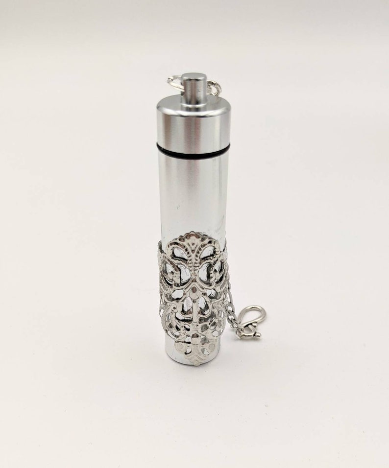 May include: A silver-colored, cylindrical pendant with a clear glass center. The pendant has a decorative filigree band and a small chain with a clasp. The top is a screw-on cap.
