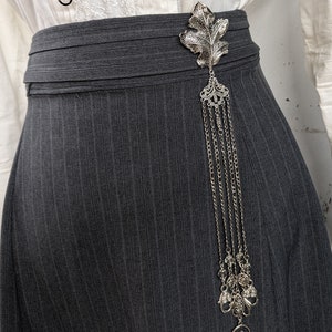 May include: A silver leaf-shaped brooch with a dangling chain of charms. The brooch is attached to a dark gray skirt.