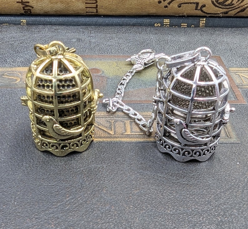May include: Two decorative birdcage pendants. One is gold-toned, the other silver-toned. Both feature a bird silhouette and ornate detailing. Each cage is suspended from a chain. The cages are likely intended for jewelry or decorative purposes.