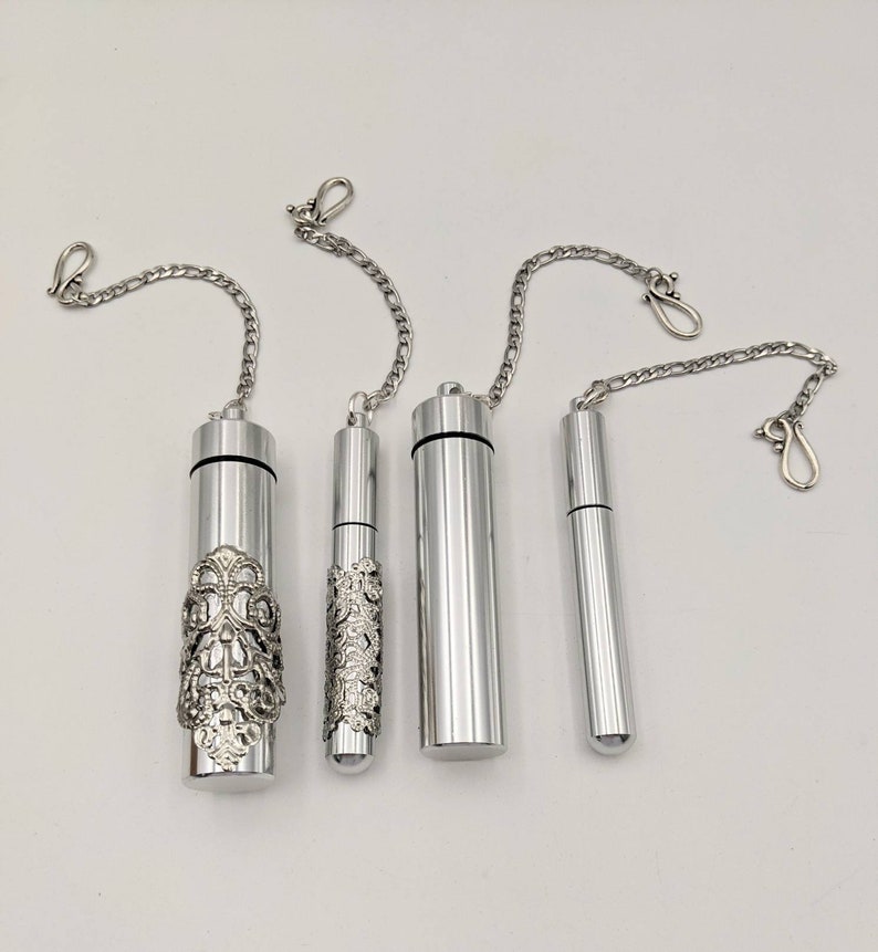 May include: Four silver-colored, cylindrical containers with attached chain necklaces. Two containers have ornate, decorative designs, while the other two are plain. Each container has a small, silver clasp at the end of the chain.