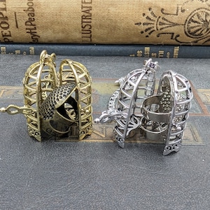 May include: Two decorative cage-shaped pendants. One is gold-toned, and the other is silver-toned. Each cage opens to reveal a detailed interior. The gold cage contains a detailed object, while the silver cage has a simple interior.