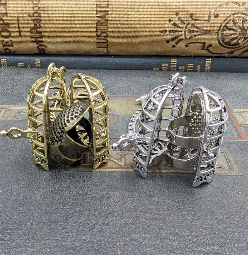 May include: Two decorative cage-shaped pendants. One is gold-toned, and the other is silver-toned. Each cage opens to reveal a detailed interior. The gold cage contains a detailed object, while the silver cage has a simple interior.