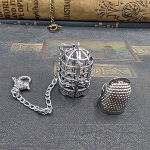 May include: A silver-colored tea infuser shaped like a birdcage, with a chain and a bird-shaped clasp. A silver thimble is also present. The items are displayed on a dark, textured surface, suggesting a vintage or antique theme.