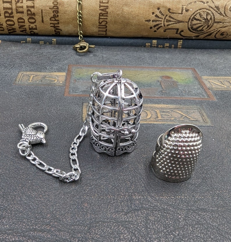 May include: A silver-colored tea infuser shaped like a birdcage, with a chain and a bird-shaped clasp. A silver thimble is also present. The items are displayed on a dark, textured surface, suggesting a vintage or antique theme.