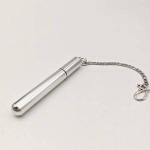 May include: A silver-colored, cylindrical container with a rounded end and a chain attached. The container has a cap and a small chain with a clasp. The item is likely a perfume or essential oil vial.
