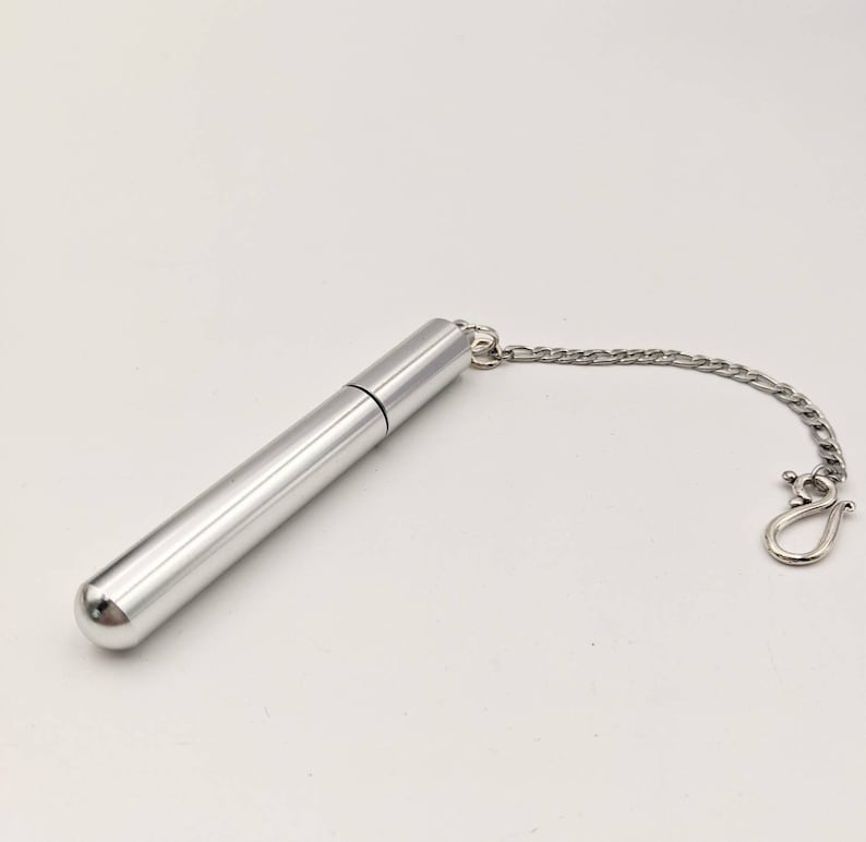 May include: A silver-colored, cylindrical container with a rounded end and a chain attached. The container has a cap and a small chain with a clasp. The item is likely a perfume or essential oil vial.