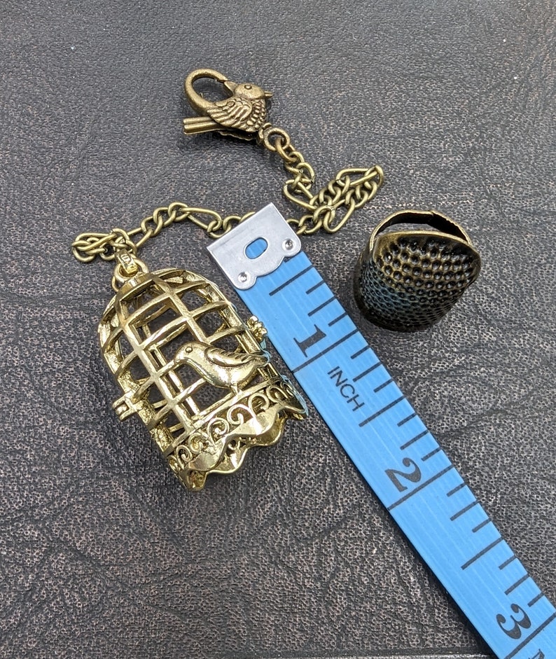 May include: A gold-colored birdcage charm with a bird inside, connected by a chain to a bird-shaped clasp. A thimble and a blue measuring tape are also present. The tape measures up to 3 inches.