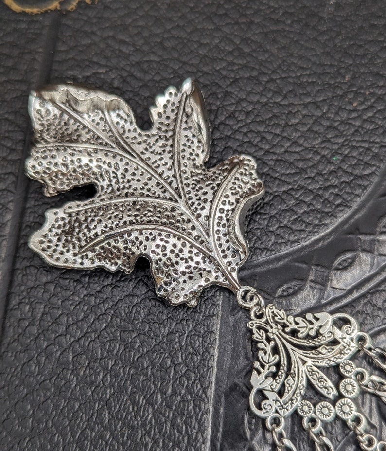 May include: Silver leaf charm with a detailed design and a dangling chain with decorative beads.