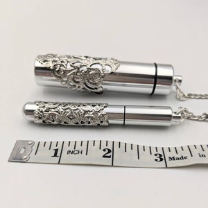 May include: Two silver-colored, cylindrical pendants with ornate filigree detailing. One pendant is larger than the other. Both pendants are attached to a silver chain. A measuring tape shows the larger pendant is approximately 3 inches long.