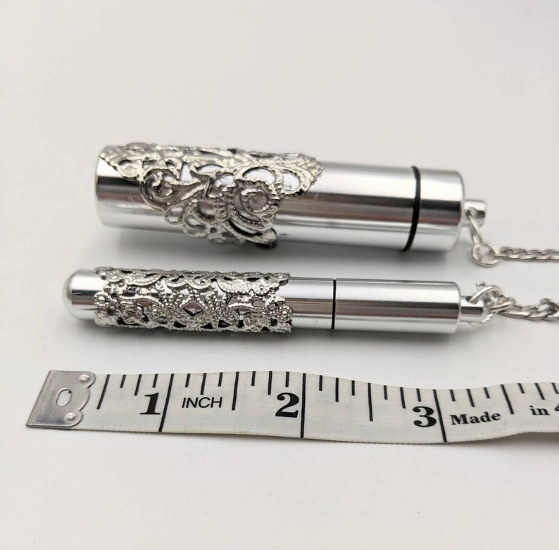 May include: Two silver-colored, cylindrical pendants with ornate filigree detailing. One pendant is larger than the other. Both pendants are attached to a silver chain. A measuring tape shows the larger pendant is approximately 3 inches long.