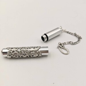 May include: Silver-colored cylindrical pendant with a decorative floral filigree design. The pendant is open, revealing the interior. A silver chain and clasp are attached, suggesting it can be worn as a necklace. The background is plain white.