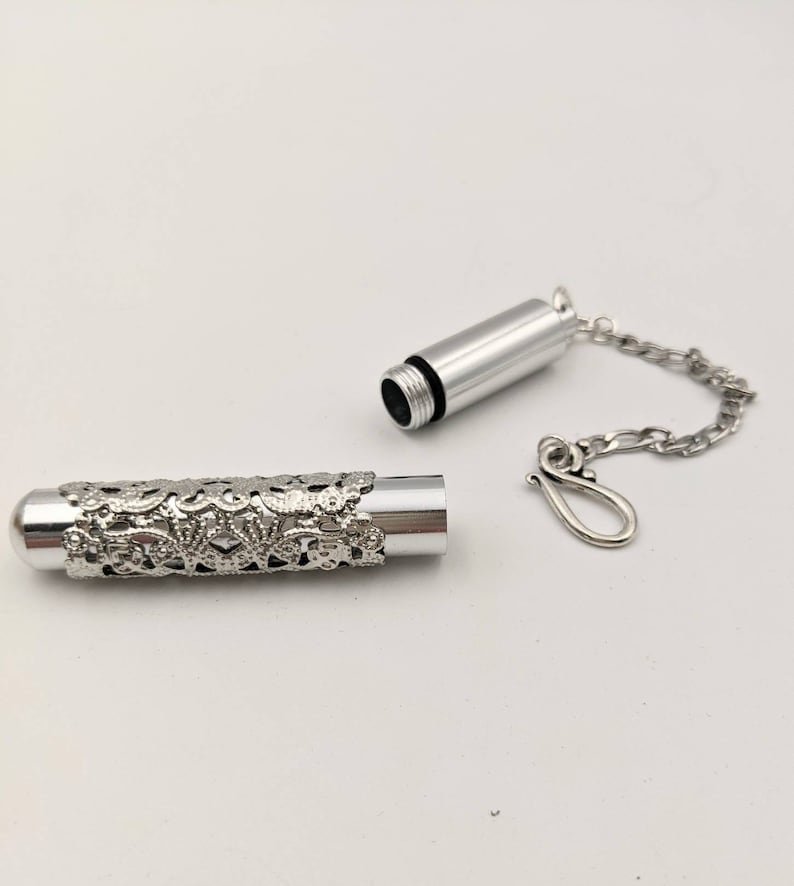 May include: Silver-colored cylindrical pendant with a decorative floral filigree design. The pendant is open, revealing the interior. A silver chain and clasp are attached, suggesting it can be worn as a necklace. The background is plain white.