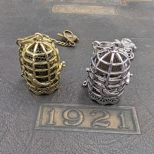 May include: Two decorative birdcage pendants, one gold-toned and one silver-toned, with attached bird charms. The cages are detailed with a bird design and are set on a dark, textured surface with the embossed year "1921".