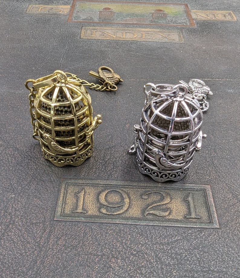 May include: Two decorative birdcage pendants, one gold-toned and one silver-toned, with attached bird charms. The cages are detailed with a bird design and are set on a dark, textured surface with the embossed year "1921".