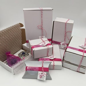 May include: Multiple white boxes with pink and white ribbon tied around them. The boxes are labeled "Razzberries Studio" and have a pink and white striped pattern. One box has a pink and white pillow with the text "Razzberries Studio" and "Hope this brightens your day!"
