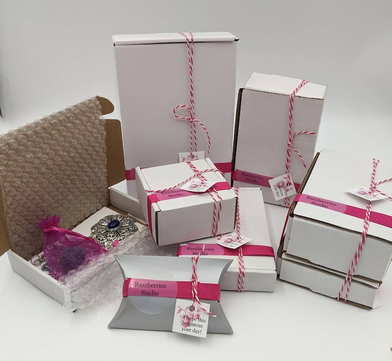 May include: Multiple white boxes with pink and white ribbon tied around them. The boxes are labeled "Razzberries Studio" and have a pink and white striped pattern. One box has a pink and white pillow with the text "Razzberries Studio" and "Hope this brightens your day!"