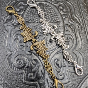 Può includere: Due ciondoli per catena decorativi con motivi floreali decorati. Un ciondolo è argentato e l'altro è dorato.