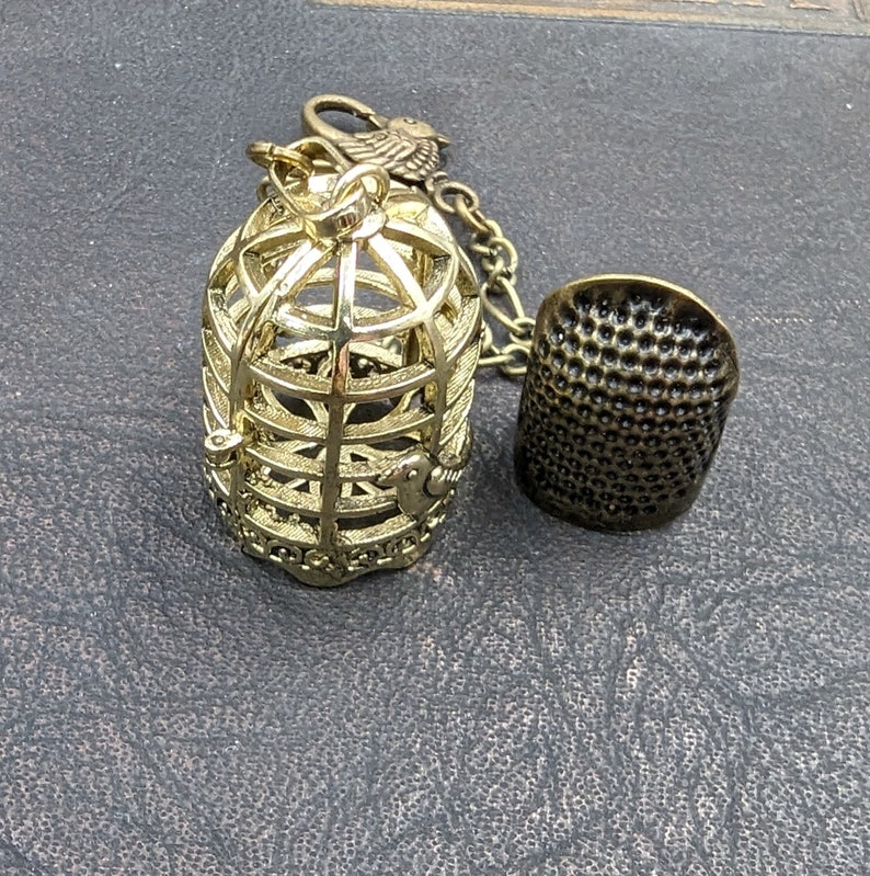 May include: A gold-colored metal birdcage charm with a small bird on top, connected by a chain to a dark bronze thimble. The thimble has a textured surface with small, evenly spaced holes. The birdcage is empty.