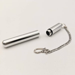 May include: A silver-colored, cylindrical container with a screw-on cap and a small chain attached. The container is open, revealing the interior. A separate, matching cylindrical piece lies next to it. The chain ends with a clasp.