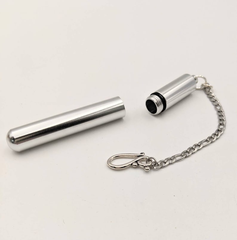 May include: A silver-colored, cylindrical container with a screw-on cap and a small chain attached. The container is open, revealing the interior. A separate, matching cylindrical piece lies next to it. The chain ends with a clasp.