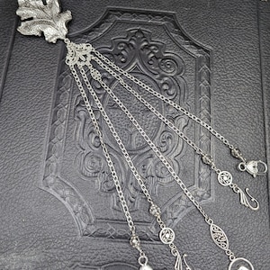 May include: Silver bookmark with a leaf design and five dangling chains. Each chain has a different charm at the end, including a bird, a heart, and a swirl.