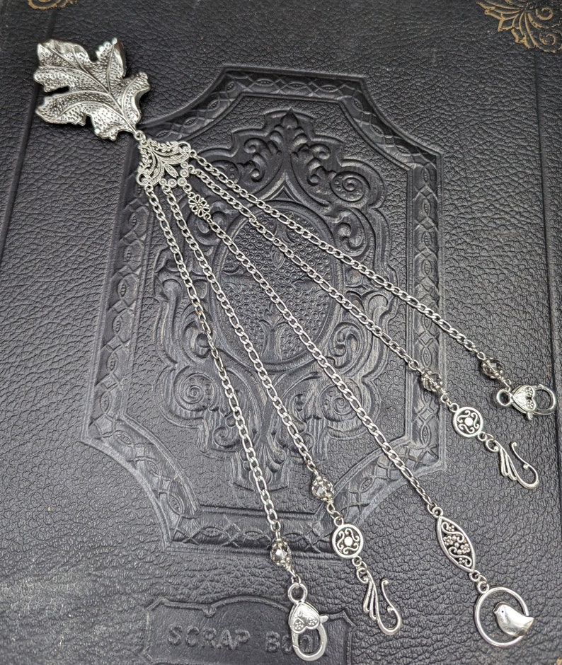 May include: Silver bookmark with a leaf design and five dangling chains. Each chain has a different charm at the end, including a bird, a heart, and a swirl.