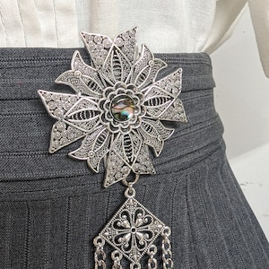 May include: A silver filigree brooch with a large iridescent shell cabochon in the center. The brooch is attached to a long chain with a square pendant.