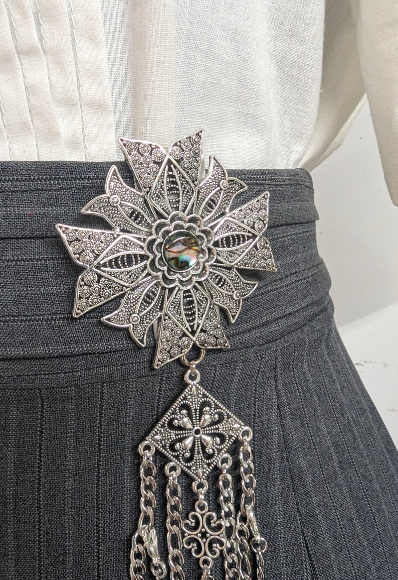 May include: A silver filigree brooch with a large iridescent shell cabochon in the center. The brooch is attached to a long chain with a square pendant.