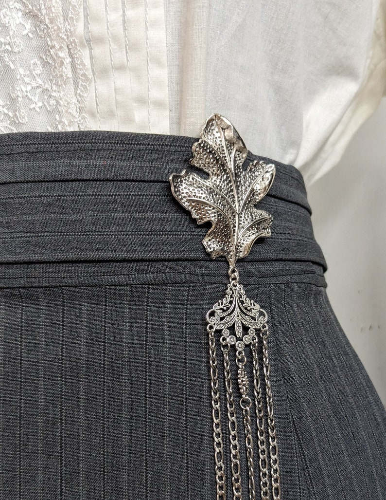 May include: A silver leaf-shaped brooch with a dangling chain of multiple strands. The brooch is attached to a gray skirt.