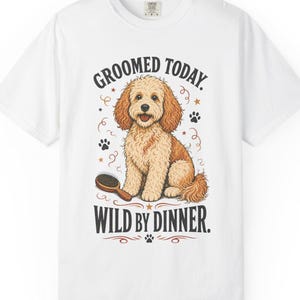 Puede incluir: Una camiseta blanca de manga corta con el gráfico de un perro de pelo rizado color marrón claro sentado junto a un cepillo. El texto sobre el perro dice GROOMED TODAY y el texto debajo dice WILD BY DINNER, decorado con huellas y estrellas.
