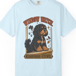 May include: A light blue t-shirt featuring a graphic of a black and tan dog sitting on a windowsill. The text reads "WINDOW WATCH." at the top and "NEIGHBOURHOOD REVIEWED." at the bottom. The design includes binoculars, paw prints, and a small bird.