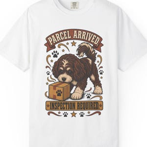 May include: A white short-sleeve t-shirt featuring a graphic of a brown and white puppy standing over a cardboard box. The text above reads PARCEL ARRIVED and the text below reads INSPECTION REQUIRED, surrounded by small stars and paw prints.