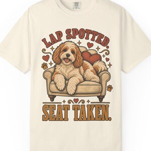 Puede incluir: Una camiseta de color crema con un gráfico de un perro marrón y blanco sentado en un sillón. El texto sobre el perro dice "LAP SPOTTED." y el texto debajo dice "SEAT TAKEN.". El diseño incluye corazones rojos y huellas marrones.