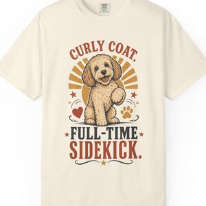 Peut inclure: Un t-shirt à manches courtes de couleur crème avec une illustration d'un chien au pelage bouclé beige. Le texte indique "CURLY COAT. FULL-TIME SIDEKICK." Le motif comprend un soleil, un cœur rouge et une empreinte de patte aux tons vintage.