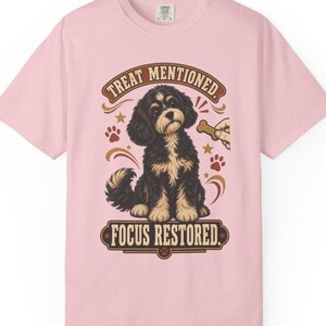 Peut inclure: Un t-shirt rose clair à manches courtes avec un motif représentant un chien noir et feu assis. Au-dessus du chien, le texte indique "TREAT MENTIONED." et en dessous, "FOCUS RESTORED.". Une main tenant une friandise est visible à droite.
