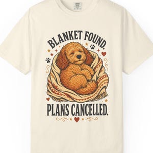 Peut inclure: Un t-shirt à manches courtes blanc cassé avec une illustration d'un chien brun doré aux poils bouclés enveloppé dans une couverture à motifs. Le texte BLANKET FOUND. PLANS CANCELLED. est imprimé en noir avec de petits cœurs rouges.