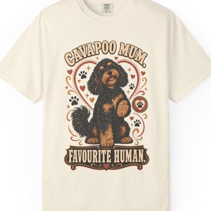 Peut inclure: Un t-shirt à manches courtes de couleur crème avec un motif représentant un chien Cavapoo noir et feu assis dans un cœur. Le texte indique CAVAPOO MUM et FAVOURITE HUMAN, entouré de petites illustrations de cœurs et d'empreintes de pattes.