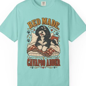 Puede incluir: Una camiseta de color turquesa claro con el gráfico de un perro Cavapoo blanco y negro tumbado en una cama con una manta de cuadros. El texto "BED MADE. CAVAPOO ADDED." aparece en una fuente retro sobre y bajo el perro, junto a huellas.