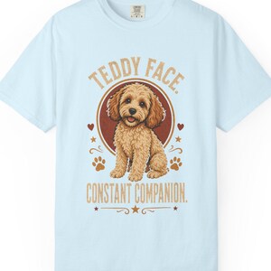 Peut inclure: Un t-shirt bleu clair avec une illustration d'un chien au pelage bouclé brun doré assis dans un cercle rouge foncé. Le texte indique TEDDY FACE. CONSTANT COMPANION. avec des empreintes de pattes, des étoiles et des cœurs autour du chien.