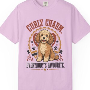 Puede incluir: Una camiseta de manga corta de color morado claro con un gráfico de un perro marrón de pelo rizado sentado dentro de un marco en forma de corazón. El texto "CURLY CHARM." aparece sobre el perro y "EVERYBODY'S FAVOURITE." debajo.