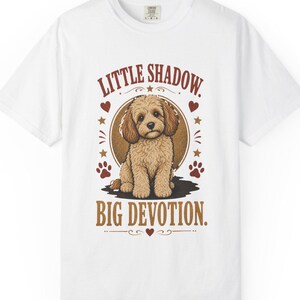 Peut inclure: Un t-shirt blanc à manches courtes avec un motif central représentant un chiot beige et poilu assis dans un cercle marron. Le texte "LITTLE SHADOW." figure au-dessus du chiot et "BIG DEVOTION." en dessous, agrémenté de petits cœurs rouges.
