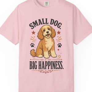 Puede incluir: Una camiseta de manga corta de color rosa claro con un gráfico de un perro de color marrón dorado y pelo rizado sentado en el centro. El texto dice SMALL DOG. BIG HAPPINESS. El diseño incluye huellas negras, estrellas rojas y líneas.