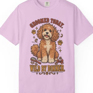 May include: A light purple short-sleeved t-shirt featuring a graphic of a brown and white curly-haired dog. The text reads GROOMED TODAY. WILD BY DINNER. The design includes a hairbrush, a bow, and paw prints surrounding the dog illustration.