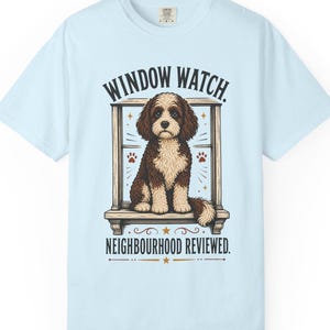 Puede incluir: Una camiseta azul claro con un gráfico de un perro marrón y blanco sentado en el marco de una ventana. El texto superior dice WINDOW WATCH. y el texto inferior dice NEIGHBOURHOOD REVIEWED. El diseño incluye huellas y estrellas pequeñas.