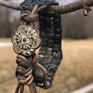 May include: A brown leather bracelet with a silver charm and black lava rock beads. The bracelet is tied with a knot and has a tassel.