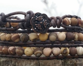May include: Wrap-around bracelet with natural stone beads in tan, brown, and cream hues. The bracelet is made with dark brown leather and a decorative copper-coloured clasp. The design wraps around the wrist multiple times.